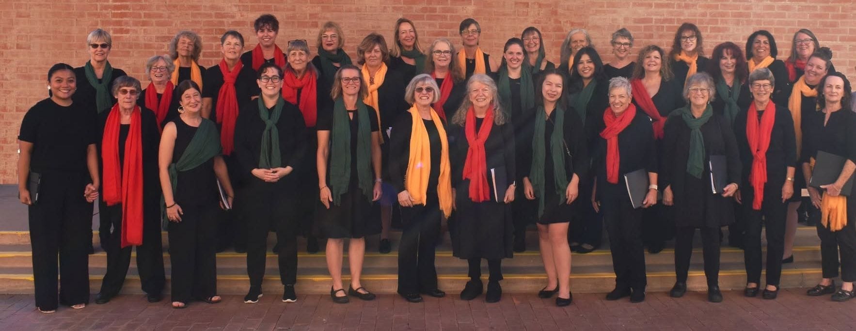 The Tucson Women's Chorus outside a brick wall, singers in black with red, green and orange scarves