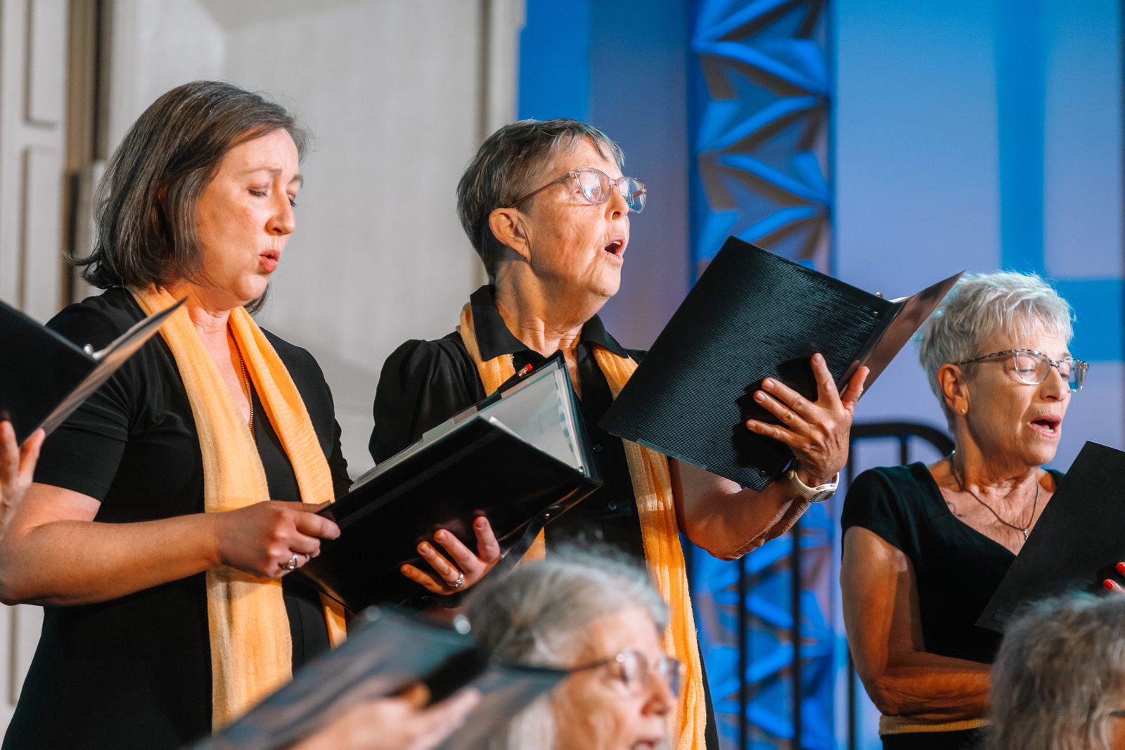 Three Tucson Women's Chorus singers holding black folders, mid-song, against a blue stage backdrop